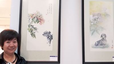 画家游淑贞与她的作品合影，图中可爱的小狗，表情生动传神。